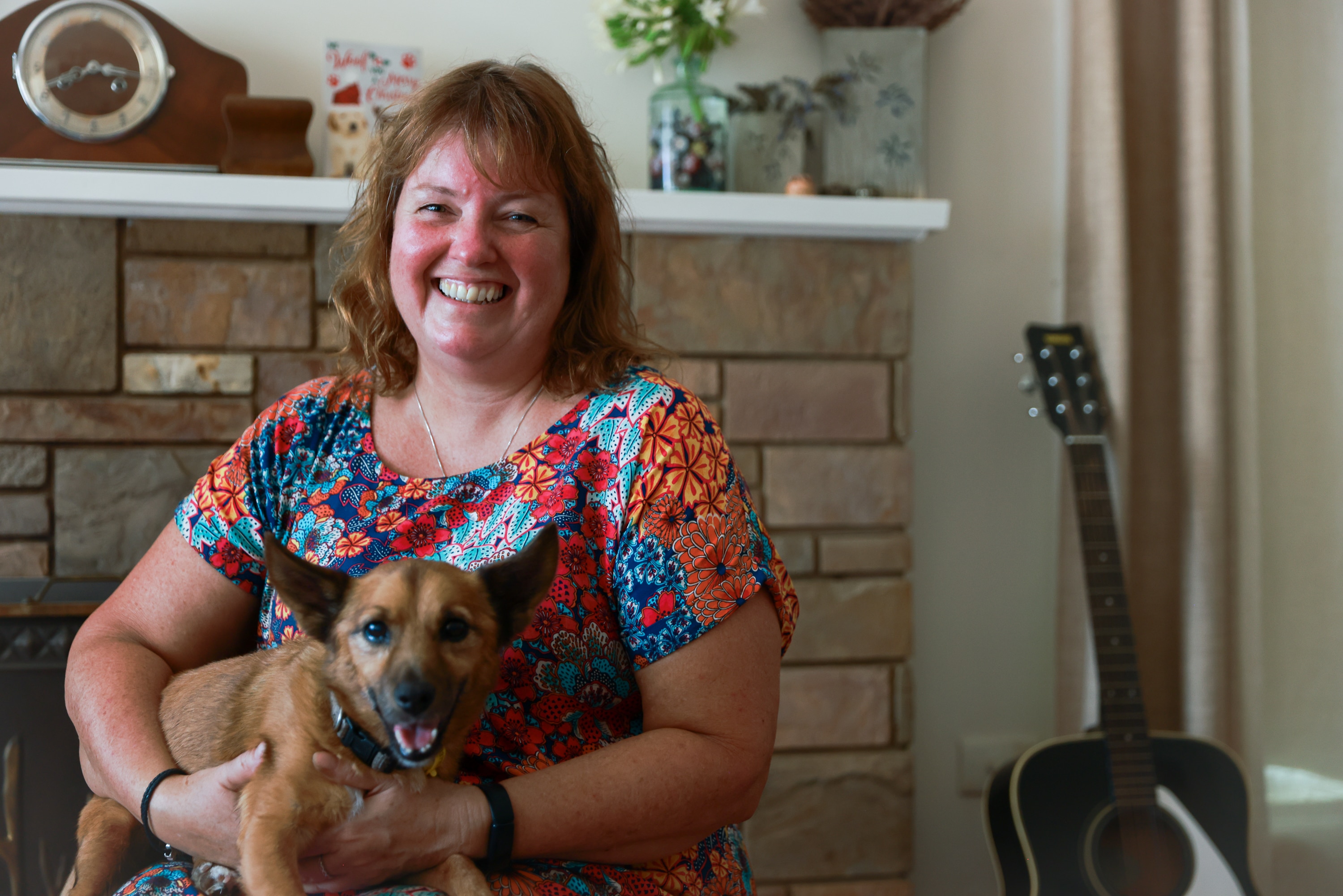A woman smiling in a colorful dress, holding her dog in a living room.