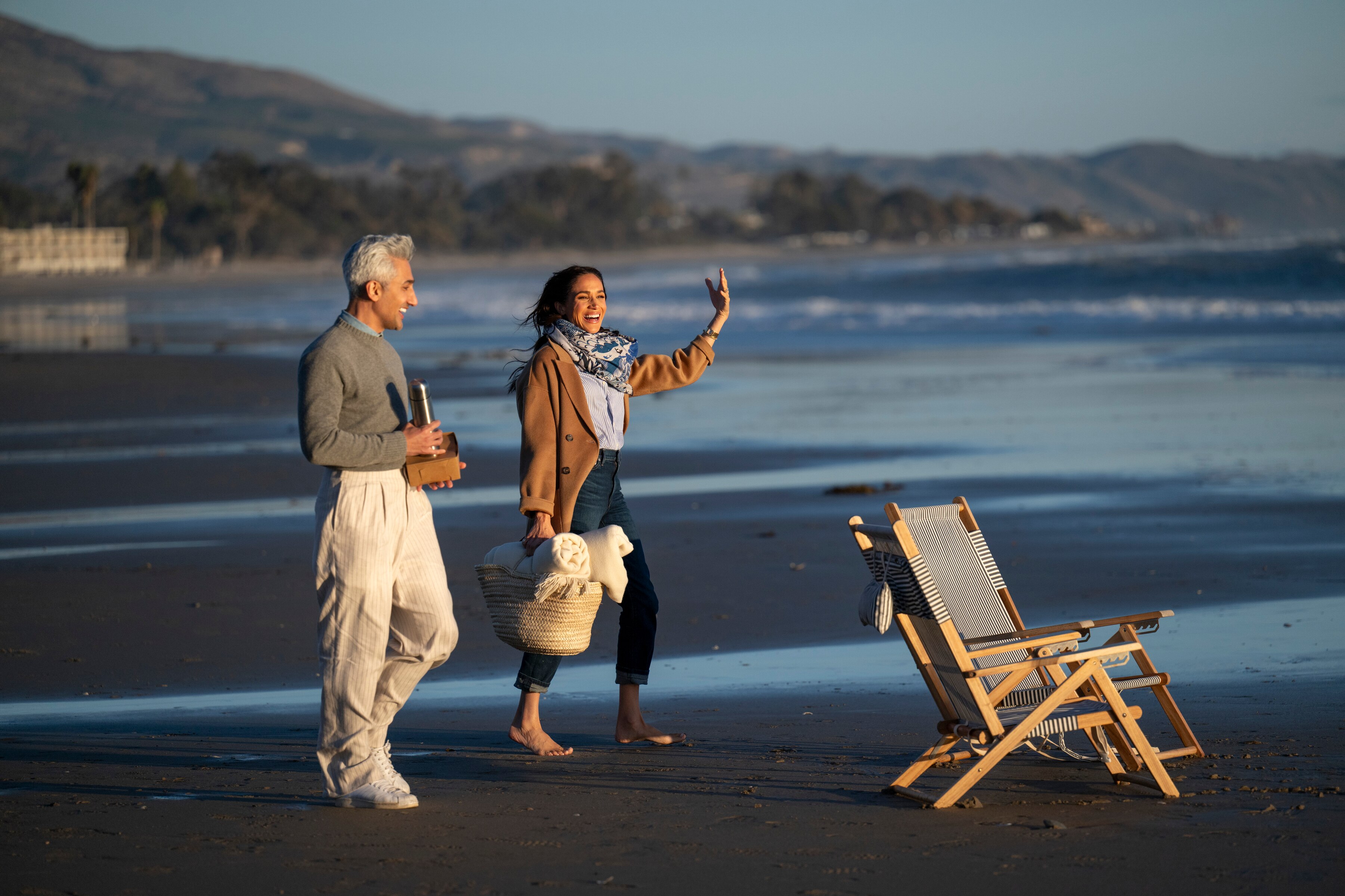 A woman & a man carrying a basket walk along a beach towards two deck chairs, they look happy and they're talking to each other