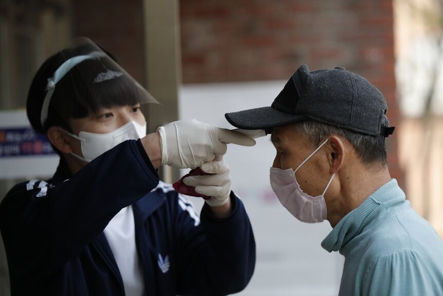 A voter in a face mask and cap gets his temperature checked by an official wearing gloves and a face shield