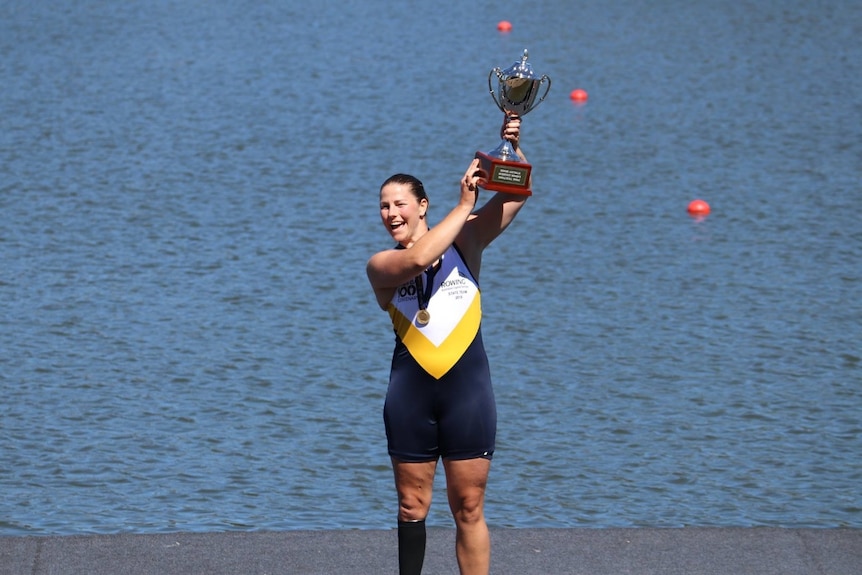 A smiling rower holds a trophy up high