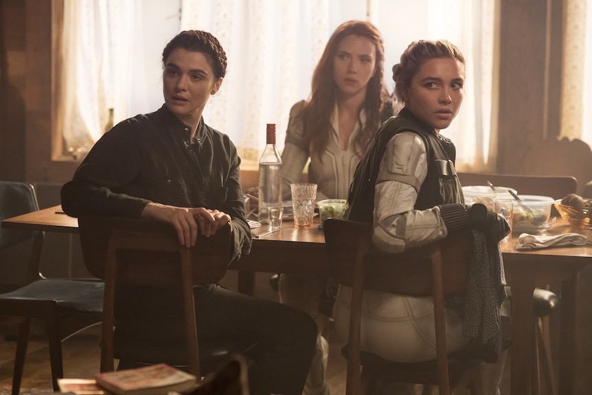 Three women sit at dinner table, turning towards camera as if looking at someone.