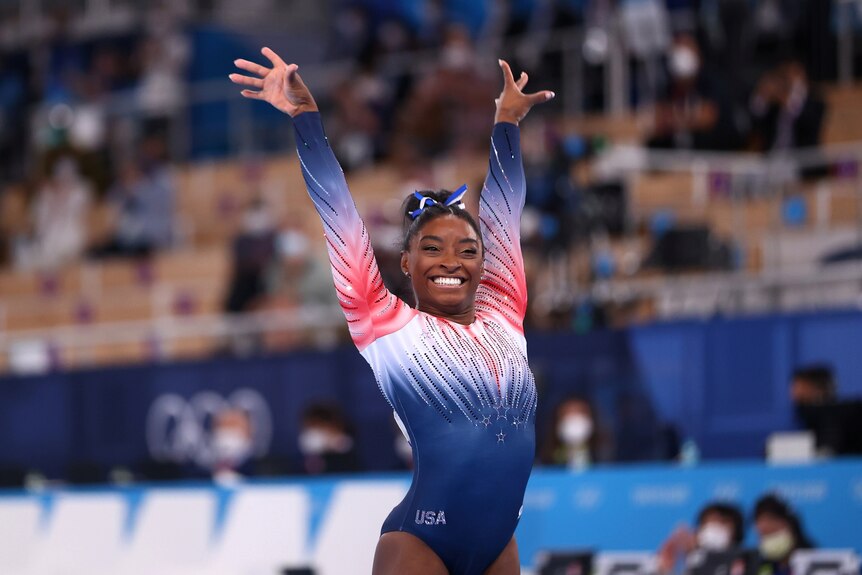 team usa gymnast simone biles finishes her routine with a big smile wearing a blue white and red leotard and a blue hair ribbon
