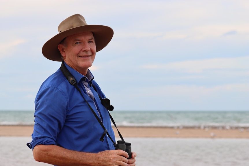 A man wearing a blue shirt and brown hat smiles at the camera. He is by the beach holding binoculars. 