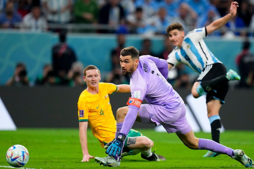 An Australian goalkeeper and defender watch the ball roll into the net as an Argentinian striker runs away in the background.