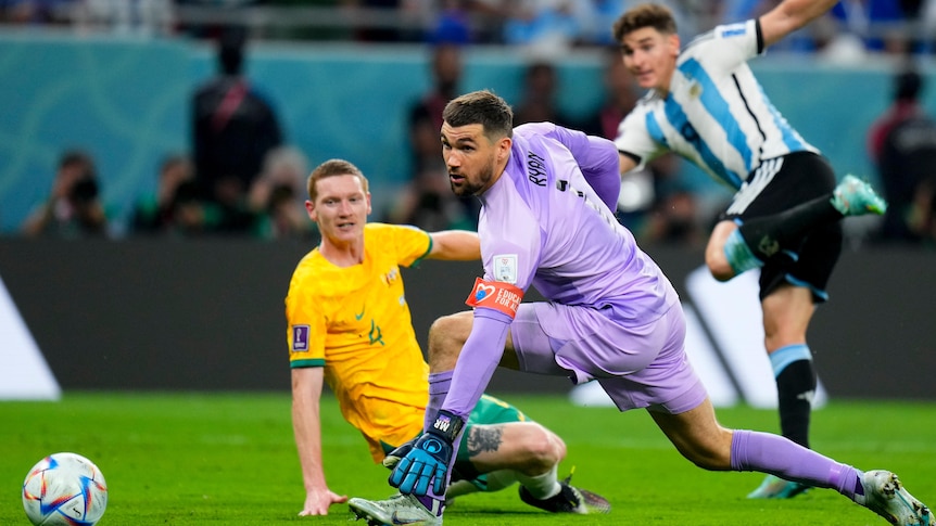 An Australian goalkeeper and defender watch the ball roll into the net as an Argentinian striker runs away in the background.