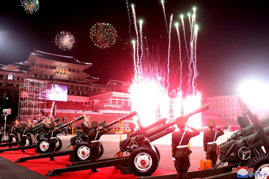 Red fireworks explode in the sky as soldiers surrounded by guns stand at attention.