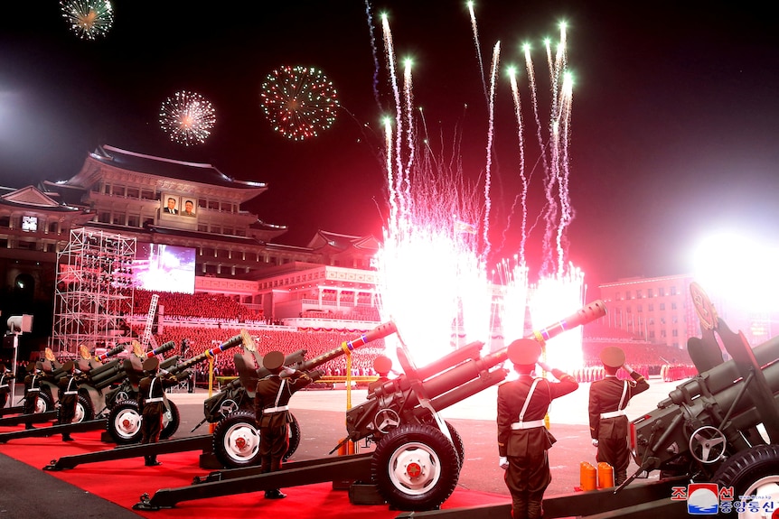 Des feux d'artifice rouges explosent dans le ciel alors que des soldats entourés d'armes à feu se tiennent au garde-à-vous.