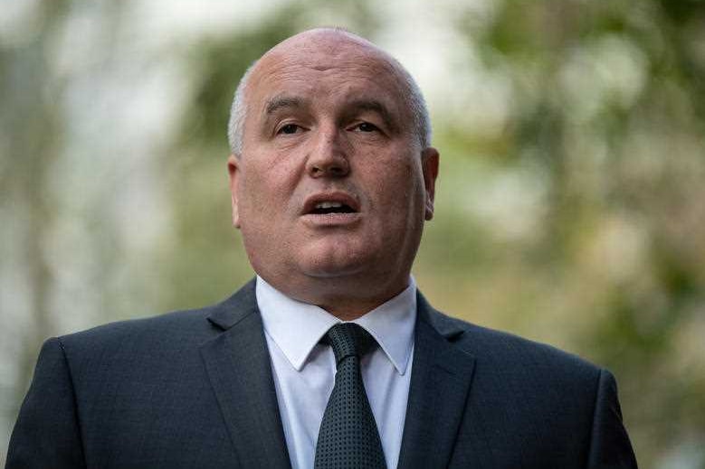 A man in a suit stands in front of foliage.