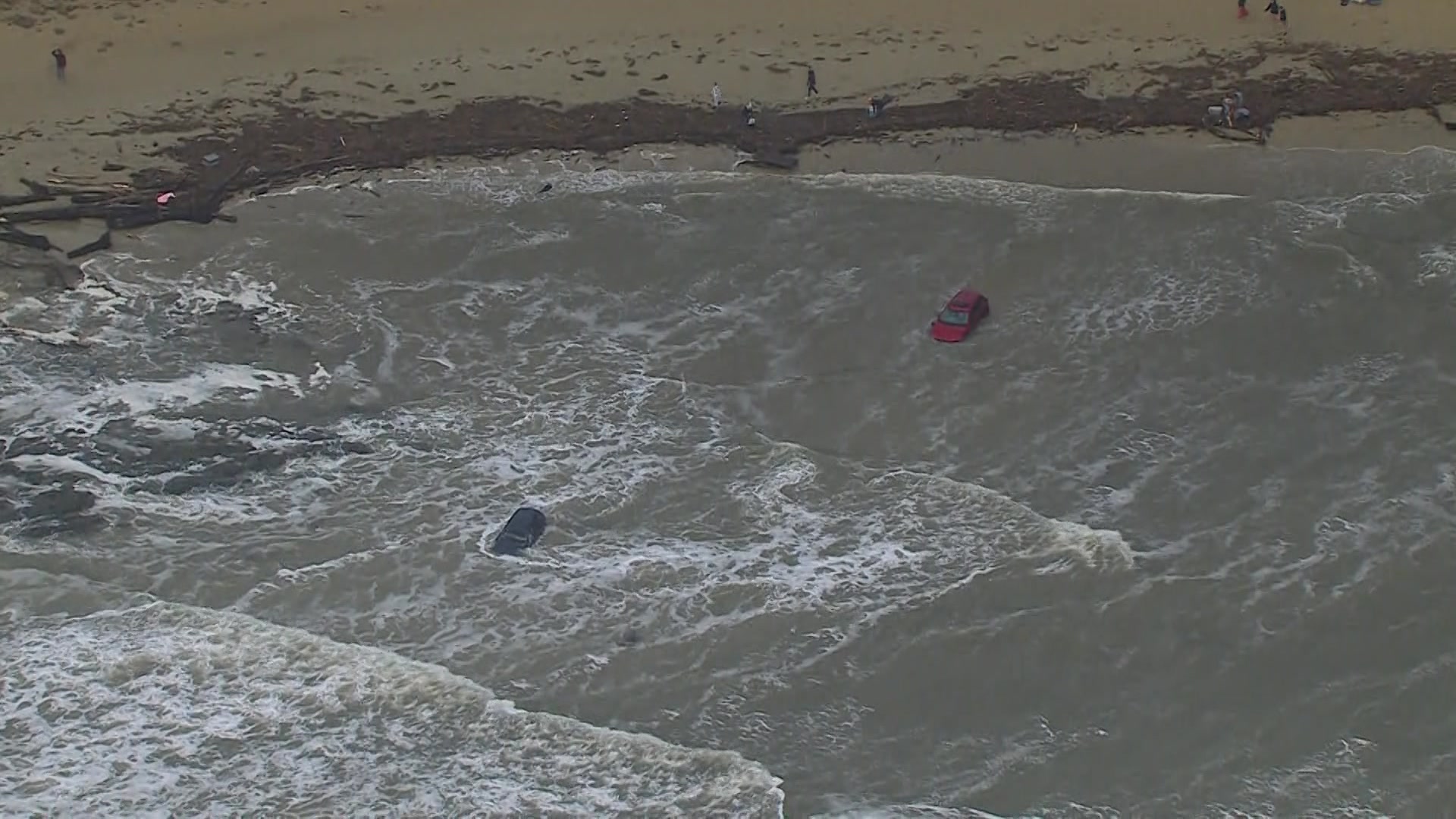 Um carro vermelho e um carro de cor escura submersos na água enquanto as ondas quebram ao redor deles perto da areia.