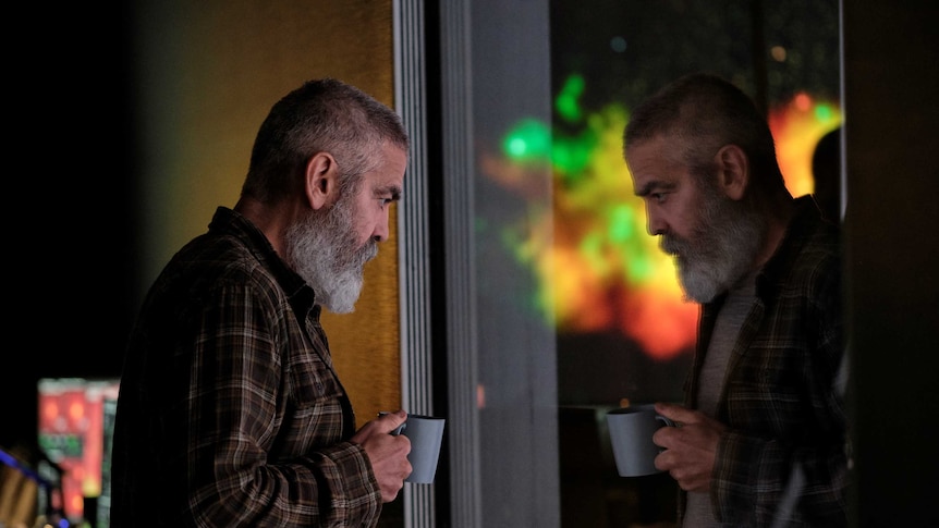 A grizzly George Clooney stares into a window with mug in hand, aurora-like light outside, in the sci-fi movie The Midnight Sky