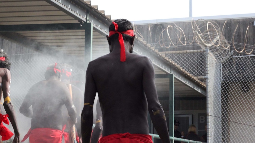 A group of traditional dressed Indigenous men surrounded by fire smoke and in front of a building.