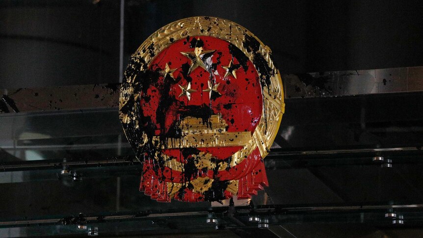 The red and gold National Emblem of the People's Republic of China is shown bolted to a glass office and is smeared in paint.