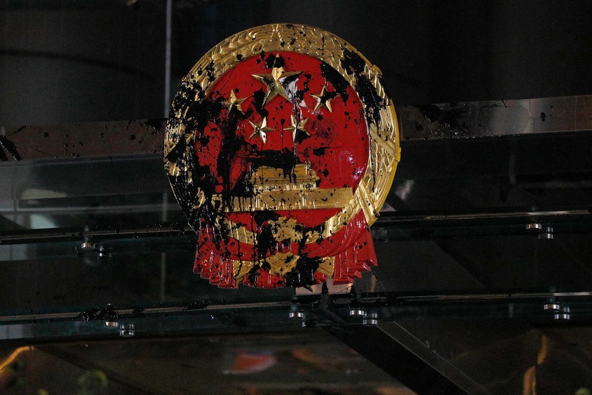 The red and gold National Emblem of the People's Republic of China is shown bolted to a glass office and is smeared in paint.
