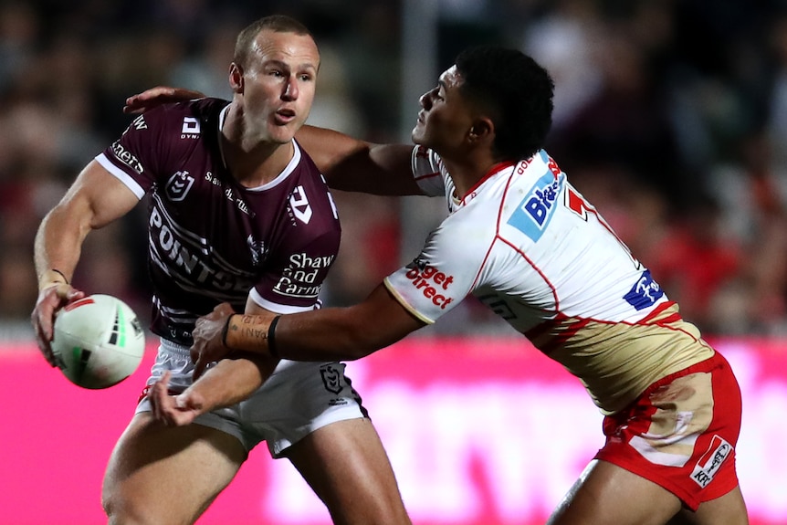 A Manly NRL player passes to his left as he is tackled by a Dolphins opponent.