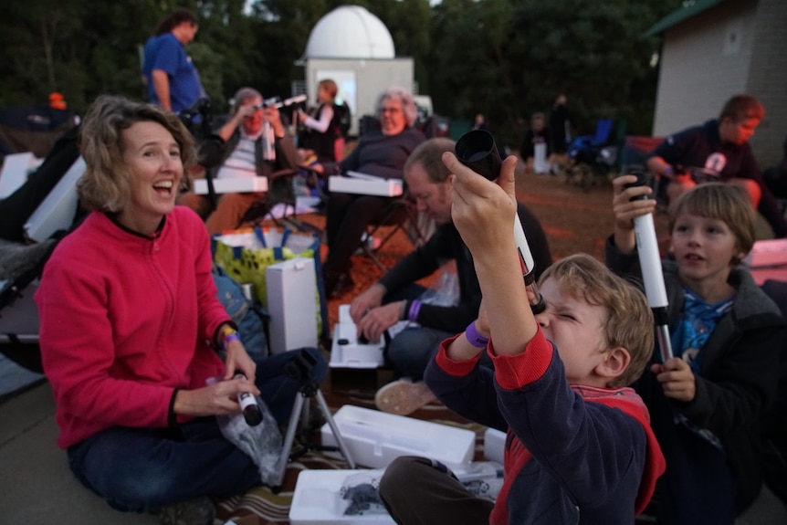 A young boy looks to the sky through a telescope while a woman smiles at him. There are more people in the background sitting.