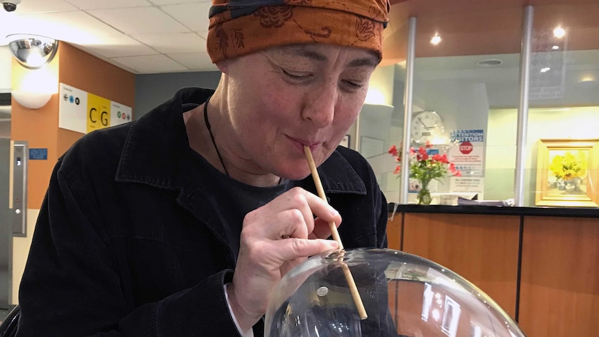 A woman in a headscarf blows air through a straw into a glass bubble in a hospital room.