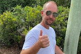 A man with short blond hair, sunglasses, and a white shirt does a thumbs up to the camera.