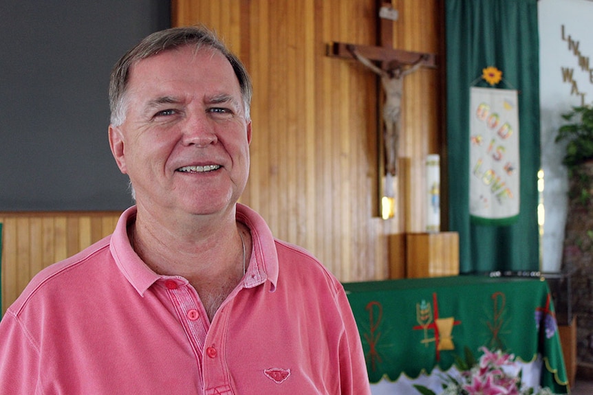 A man stands in front of a church altar with a crucifix hung on the wall behind it.