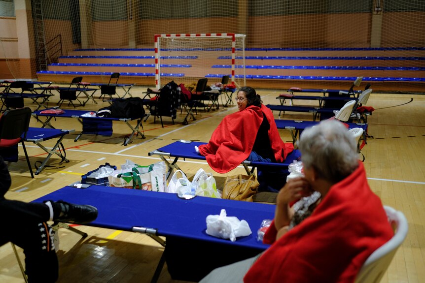 Two people wearing blankets sit in a n evacuation centre in a gym