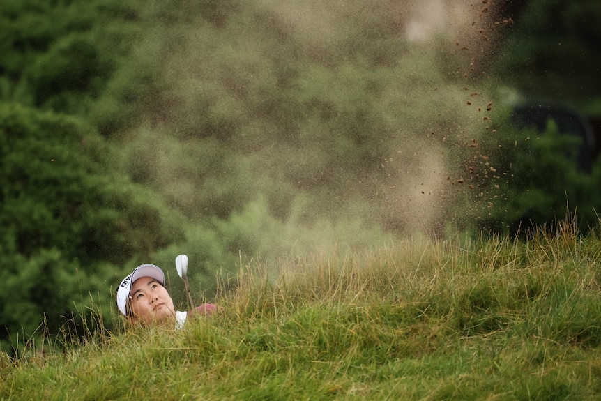 Su Oh looks out from behind a mound of grass with a puff of sand spraying with her