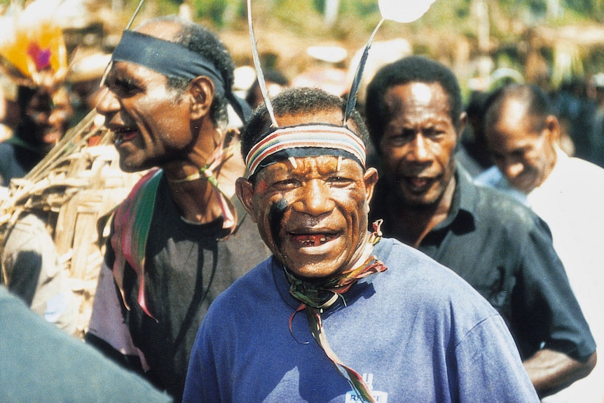 Man with black paint on face and traditional head gear marches in PNG. He is surrounded by other villagers in sea of faces. 