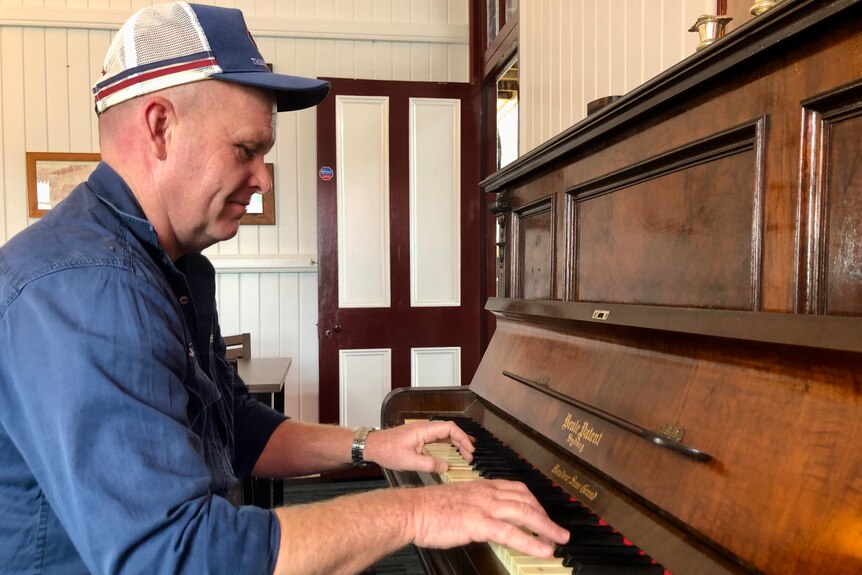A man in a cap, with a blue work shirt sits and plays a piano.
