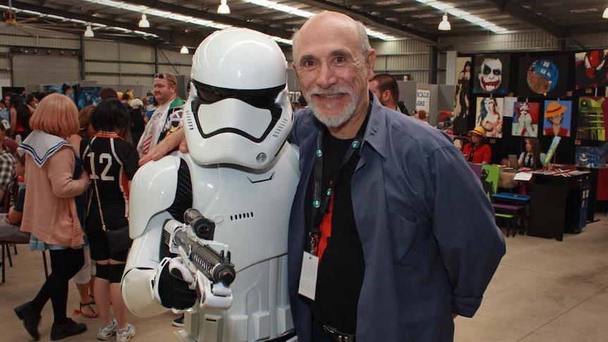 One man in a storm trooper outfit and an older gentleman with a beard smile in front of a crowd.