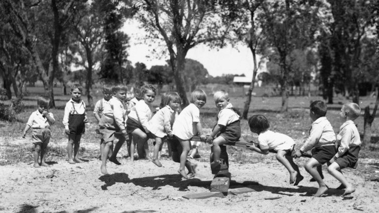 A black-and-white photo shows a group of several children playing on a see-saw.