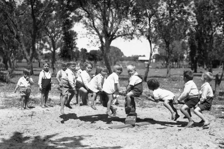 A black-and-white photo shows a group of several children playing on a see-saw.