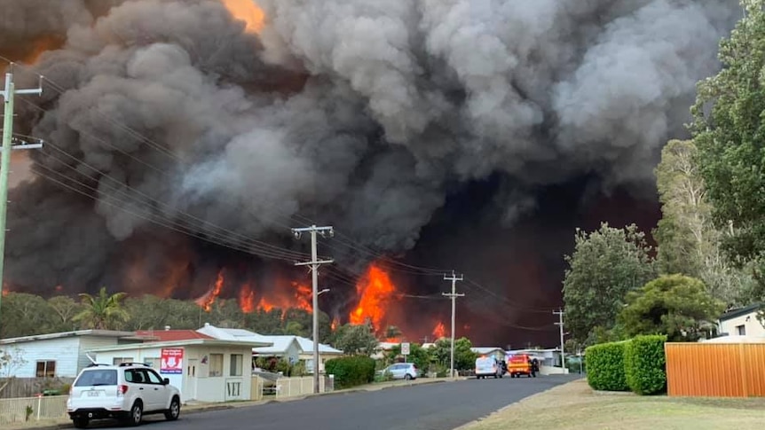 thick black clouds of smoke billow above red flames behind houses on a suburban street.