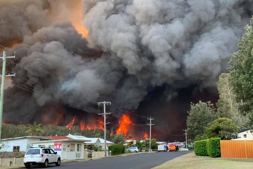 thick black clouds of smoke billow above red flames behind houses on a suburban street.