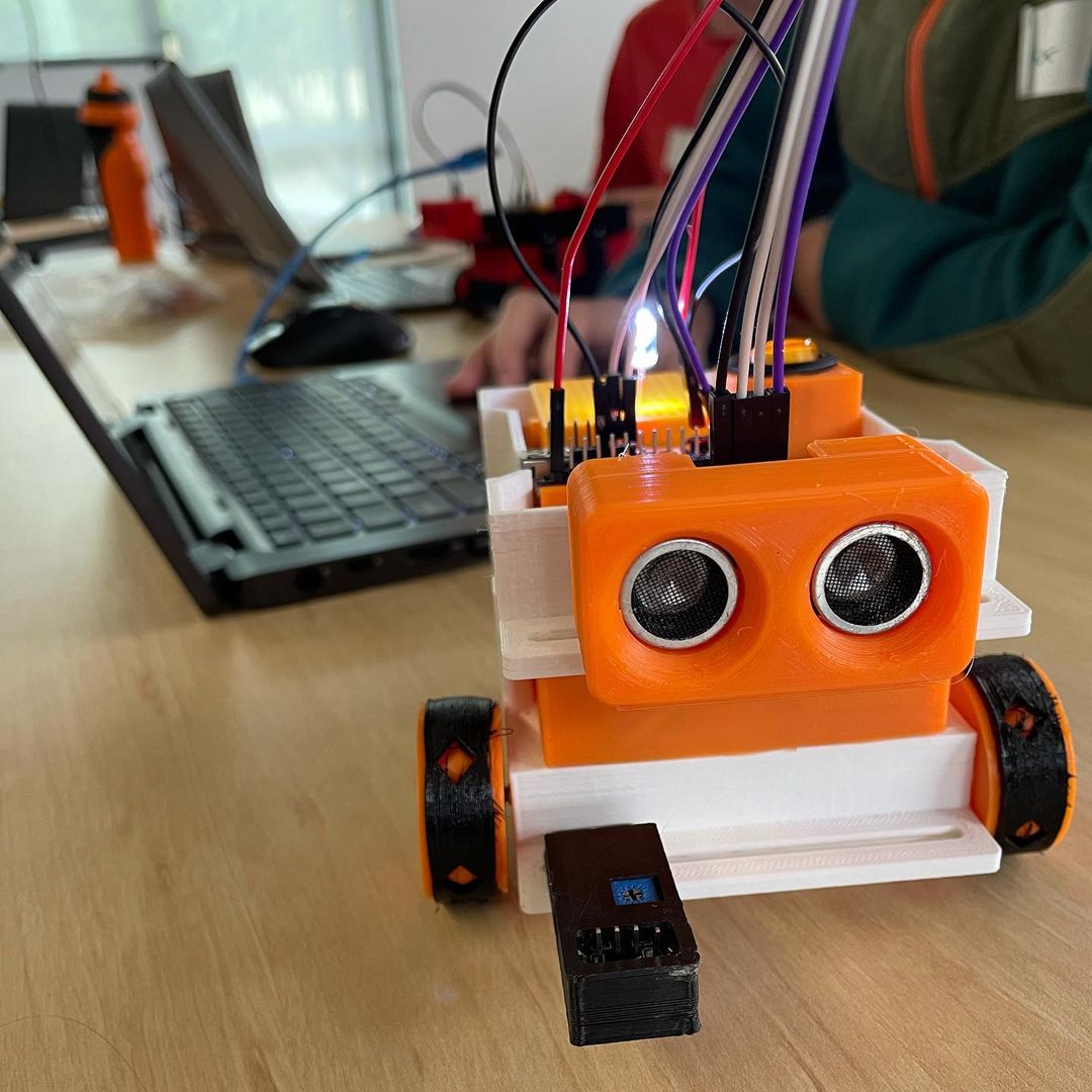 A small lego robot with wires is connected to a laptop in a classroom.