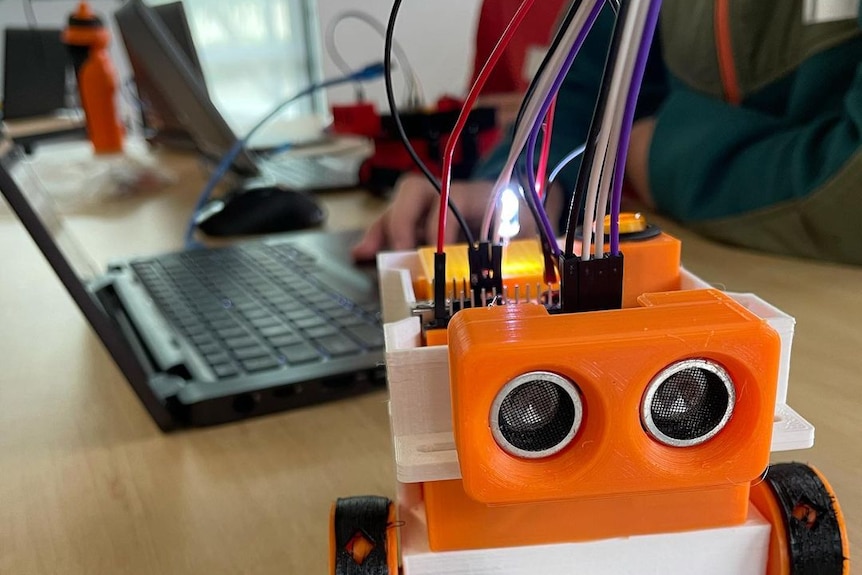 A small lego robot with wires is connected to a laptop in a classroom.