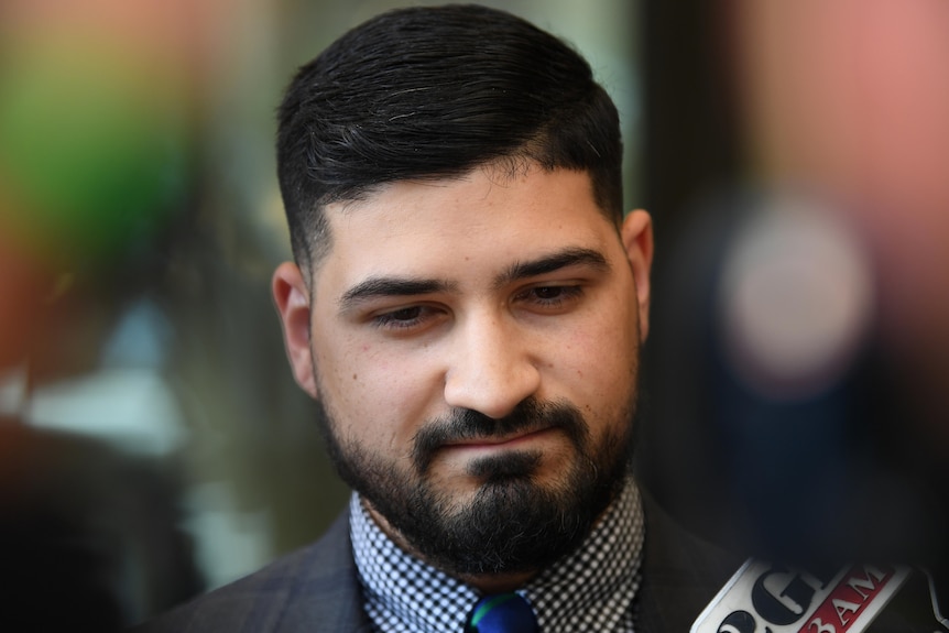 A man wearing a suit and tie looks down as a microphone is held to his face