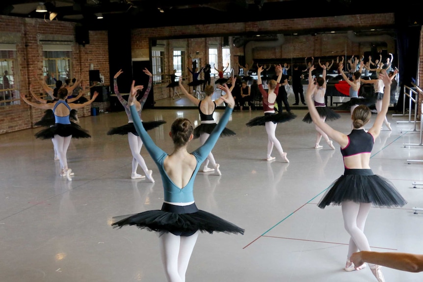 a dozen dancers stand in a mirrored studio with their arms in the air.