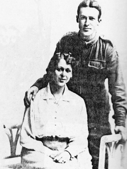 A man in a uniform stands beside a woman sitting down.