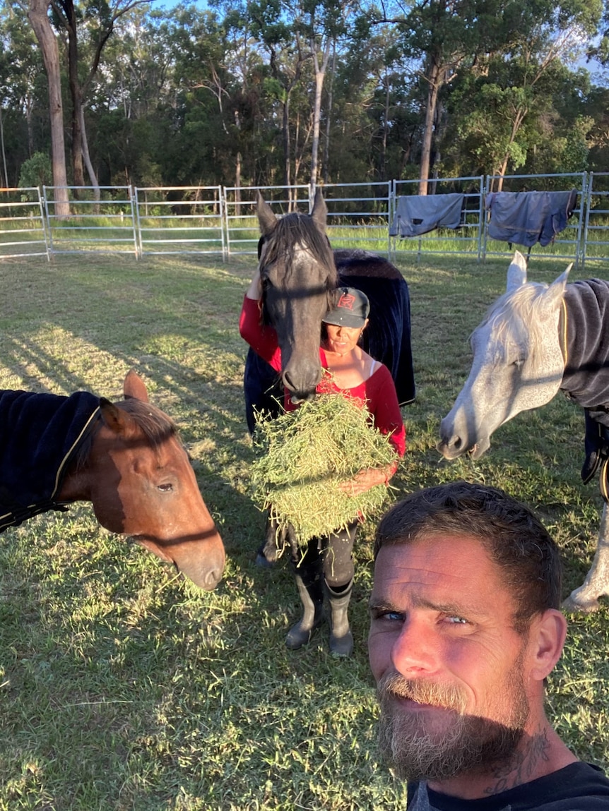 A man takes a selfie as a woman surrounded by horses, hugs and feeds a horse.