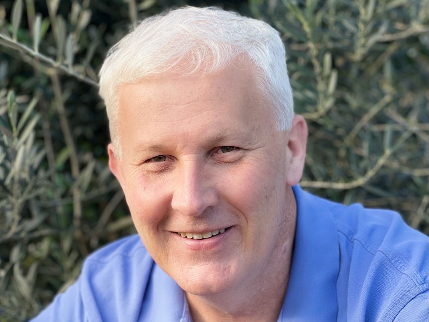 A man with white hair and a blue shirt smiles at the camera. Olive trees are in the background