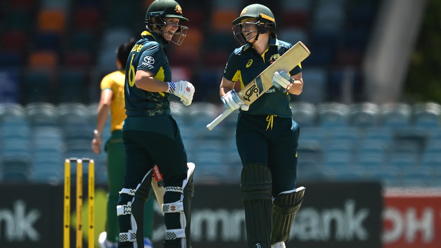 Two female crickets, playing for Australia, talk between overs, holding their bats, wearing helmets