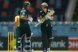 Two female crickets, playing for Australia, talk between overs, holding their bats, wearing helmets