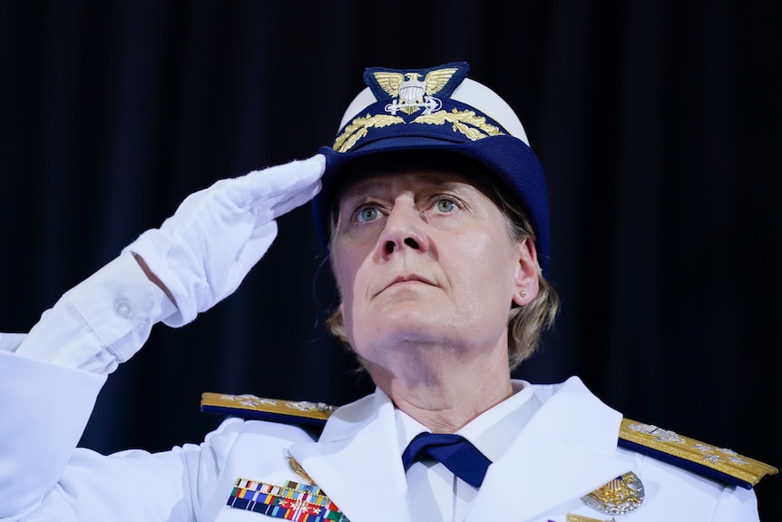 A woman dressed in white, with gold lapels, and wearing white gloves and a blue, white and gold hat, is seen saluting.