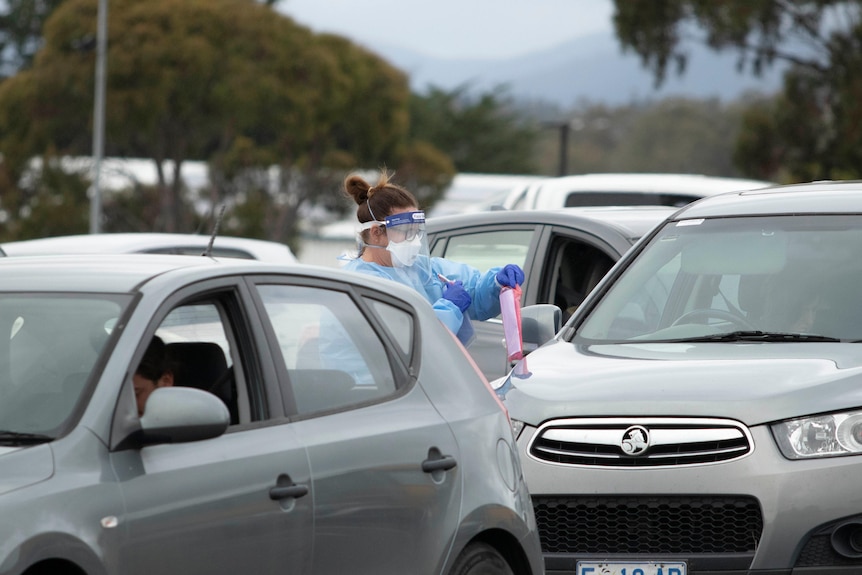 A woman in protective clothing holds a pink bag outside a car window