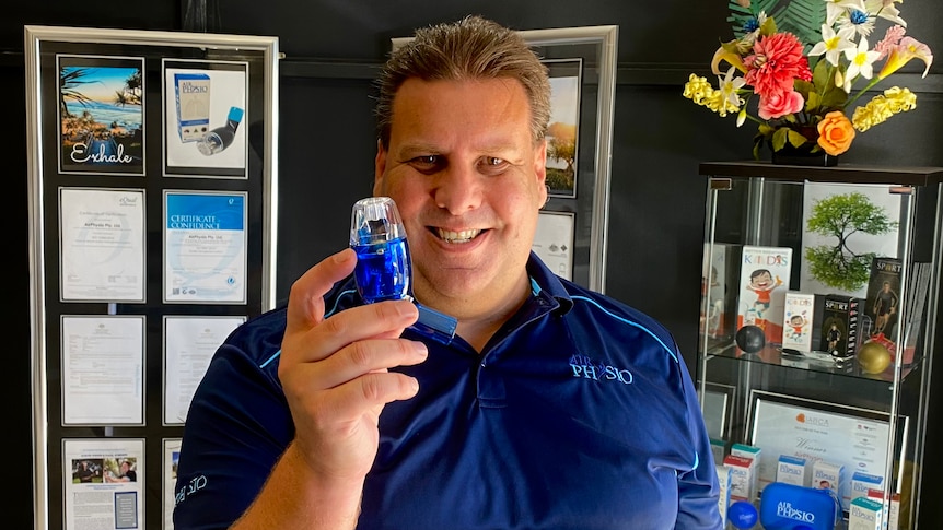 A man wearing a bright blue polo shirt holdinhg a small breathing device in his hand smiles at the camera.