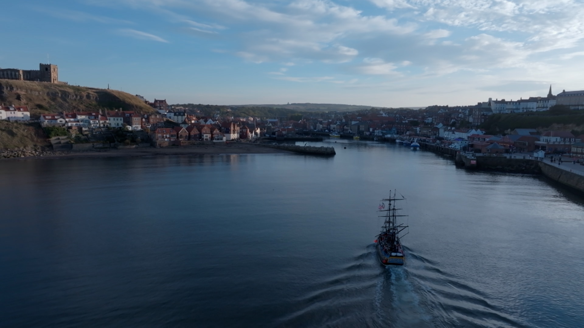 The port of Whitby with a boat sailing into it on a sunny day with a few clouds.