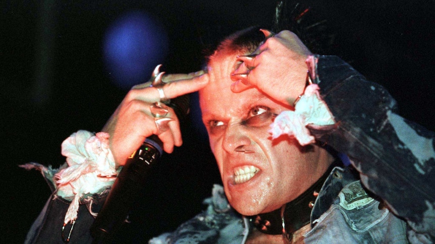 Keith Flint on stage wearing a denim jacket and lots of silver spiky rings and a spiked black collar