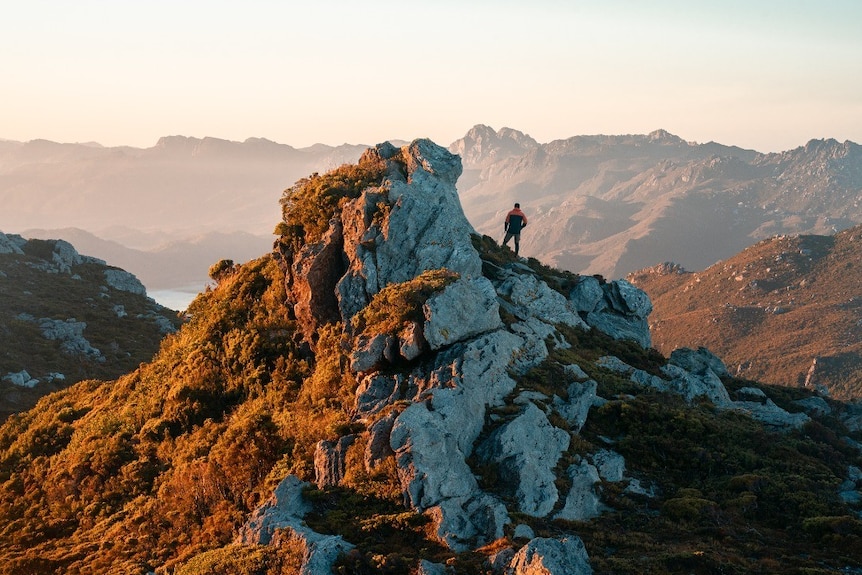 A man stands facing away from camera on a rocky outcrop, in background is water and other mountains