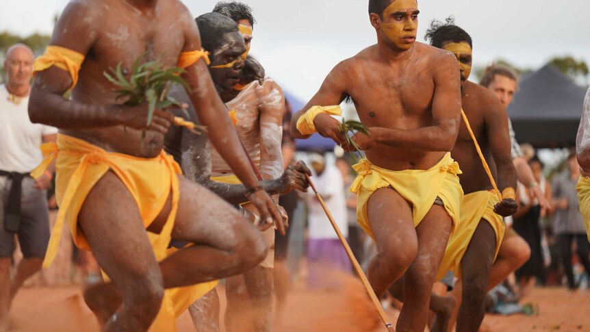 Michael Yunupingu and other Gumatj men, perform in red sand, as a crowd watches on.
