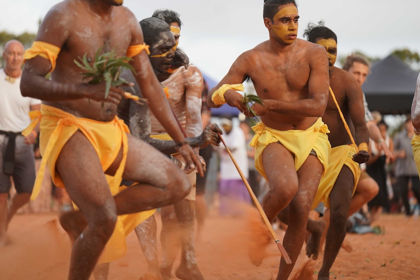 Michael Yunupingu and other Gumatj men, perform in red sand, as a crowd watches on.