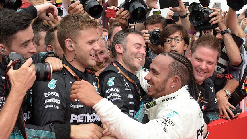 Lewis Hamilton smiles with his pit crew with camera lenses pointing down on him from behind his team