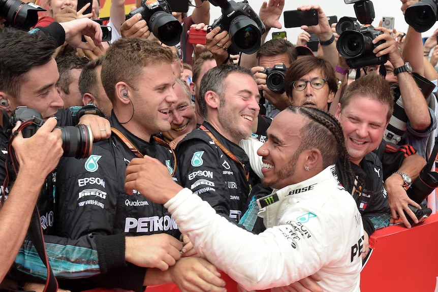 Lewis Hamilton smiles with his pit crew with camera lenses pointing down on him from behind his team
